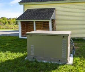 A residential standby generator sits on a concrete pad beside a house and storage shed in a grassy yard.