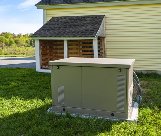 A residential standby generator sits on a concrete pad beside a house and storage shed in a grassy yard.